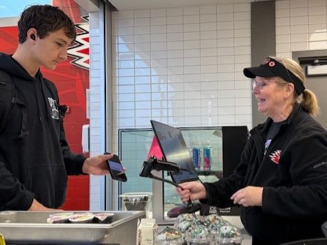 A cashier assisting a young man at a checkout counter, with the young man holding a smartphone and the cashier using a POS system.