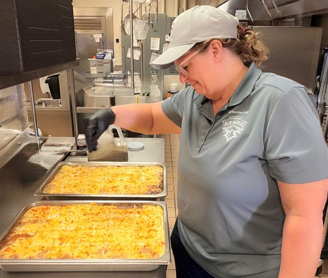 A woman in a gray shirt and cap is seasoning trays of baked pasta in a commercial kitchen.