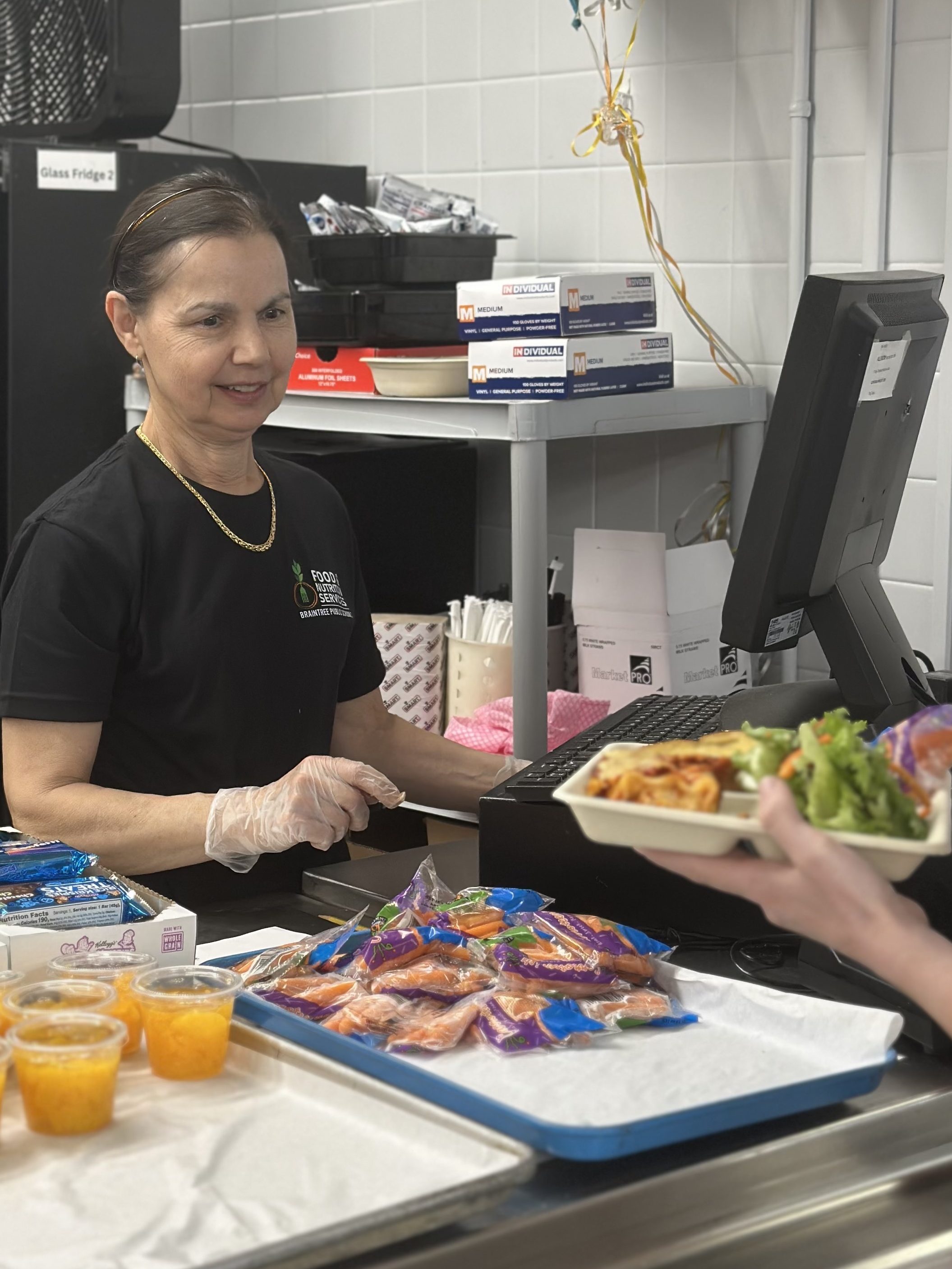 A woman in a black shirt smiles while assisting a customer at a food service counter, surrounded by packaged snacks, fruit cups, and fresh salad.