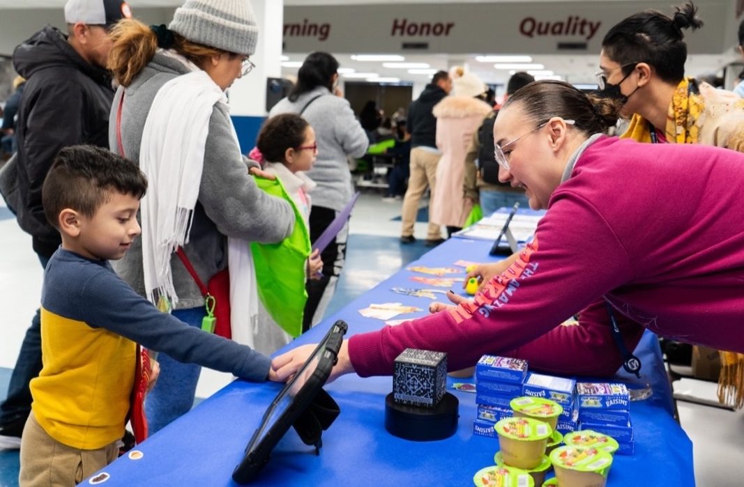 A young boy interacts with a woman at a table displaying educational materials and snacks, in a busy community event setting.