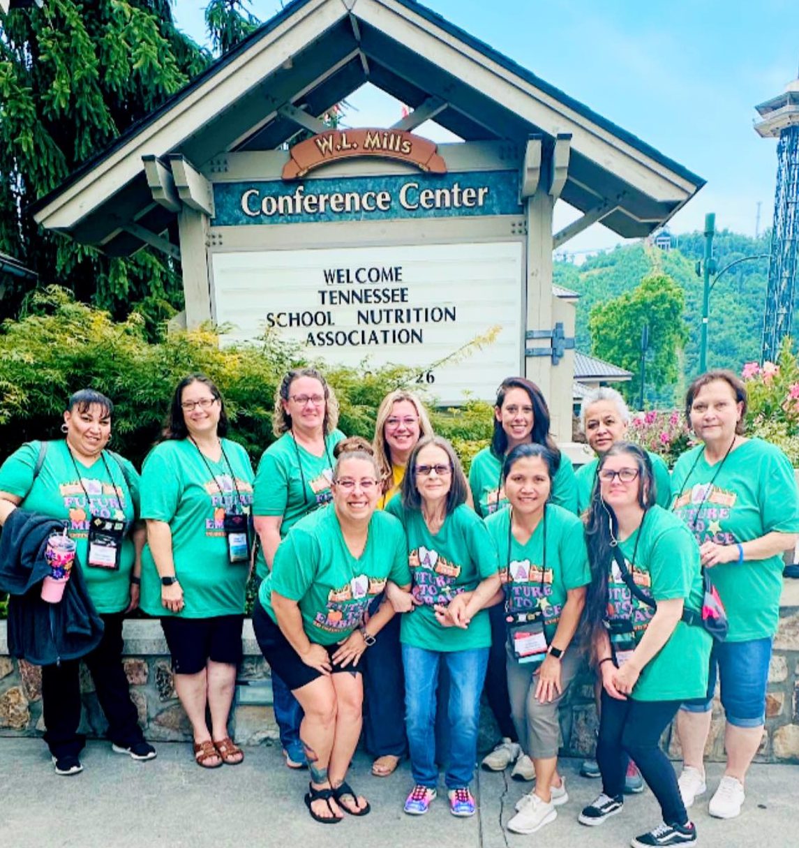 Group of ten women wearing matching green shirts posing in front of the W.L. Mills Conference Center sign welcoming the Tennessee School Nutrition Association.